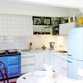 На фото: оборудованная и меблированная кухня-столовая, плита, мойка, холодильник, стенные навесные шкафы, напольные тумбы-столы, стены облицованы плиткой, на переднем плане - сервированный обеденный стол белого цвета со стульями