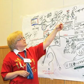 На фото: женщина в красной футболке с надписью ЦАРЬ стоит с указкой у плаката со схемой и что-то объясняет, смотрит на схему