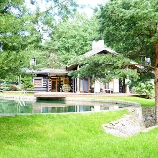 На фото: одноэтажный загородный дом из бревнав окружении сосен, перед домом - открытый бассейн, на переднем плане - газон