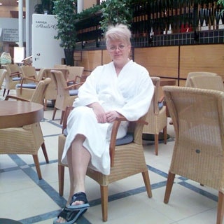 На фото: женщина в белом банном халате и шлепанцах, сидящая в кресле в холле, меблированным типовыми столиками и креслами