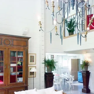 На фото: светлое внутреннее помещение, на переднем плане - шкаф, галерея второго этажа с перилами, кованая потолочная многорожковая люстра, на заднем плане - цветы на высоких подставках, обеденный стол со стульями