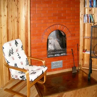 На фото: печь - камин, обложенная кирпичом, слева от печи - кресло, справа - книжный стеллаж с книгами, полы - дощатые, стены обшиты вагонкой
