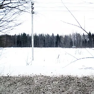 На зимнем фото: обширный земельный участок под снегом от дороги до опушки леса на заднем плане, на участке - столб низковольтной линии электропередач, участок свободен от деревьев, кустов и построек