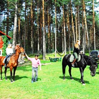 На летнем фото: на травяном газоне участка - две девушки верхом на лошадях, одну лошадь держит под уздцы мужчина, на заднем плане - телега со скошенной травой, опушка соснового леса и двухэтажная бревенчатая постройка со скатной крышей