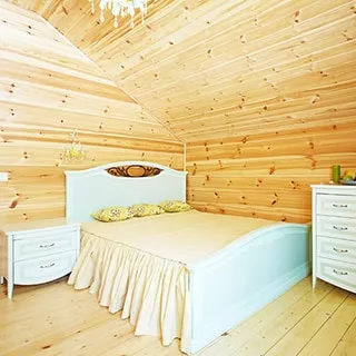 На фото: часть жилого помещения - спальни, в углу - большая двуспальная кровать с высоким деревянным изголовьем, слева от кровати - тумбочка. справа - комод, в изголовье кровати на стене - бра, на потолке люстра, пол - дощатый, стены - брус, разновысокий потолок обшит вагонкой