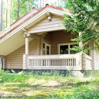 На летнем фото: одноэтажный деревянный дом с террасой и скатной кровлей, окна и входная дверь - стеклопакеты, перед домом - газон, на участке - хвойные деревья