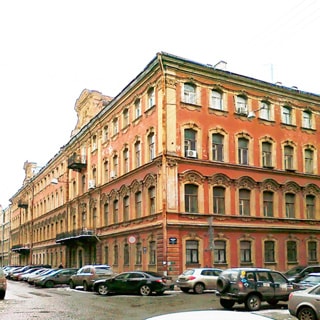 На фото: часть фасада 4‑этажного здания старой постройки, вид с угла здания, проезжая часть прилегающих улиц с припаркованными автомобилями