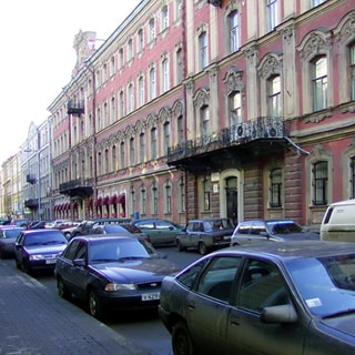 На фото: часть фасада 4-этажного здания старой постройки, с балконом, тротуар и проезжая часть прилегающей улицы с припаркованными автомобилями