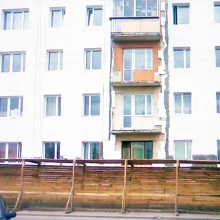 На фото: часть фасада многоэтажного (в кадре видны первые четыре этажа) многоквартирного жилого дома, на фасаде производятся ремонтные работы, на пешеходном тротуаре установлено защитное деревянное ограждение для пешеходов