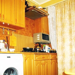 На фото: часть помещения кухни, окно закрыто шторой, от окна вдоль стены - кухонный гарнитур, столы-тумбы с общей столешницей, газовая плита, над ней - вытяжка, стиральная машина, навесные кухонные шкафы, микроволновая печь