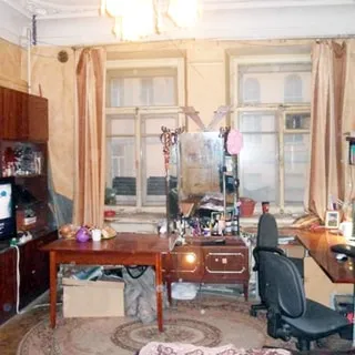 На фото: часть помещения жилой комнаты, два окна, деревянные рамы, у окна - туалетный столик с большим зеркалом, слева у стены - сервант, справа у стены - письменный стол и офисный стул, полы - линолеум, стены оклеены обоями, на потолке - люстра