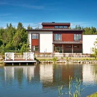 На фото: современный двухэтажный загородный дом в стиле скандинавского конструктивизма, на переднем плане - пруд с оборудованным выходом к воде, на заднем плане за домом - лесной массив