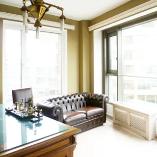 На фото: часть помещения жилой комнаты - кабинета, два окна, в углу - мягкий кожаный диван, слева от него - стул, еще левее - деревянный письменный стол, на столе - стекло, на стелке - письменные приборы, стены - окрашены, на потолке - люстра