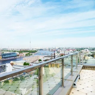 На фото: панорамный вид на город с огороженной площадки на крыше дома, река, городские кварталы по обеим берегам реки, впереди вдалеке - телевизионная башня, ограждение площадки - металло-стеклянное, полы - плитка