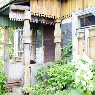 На фото: часть фасада дома, крыльцо с козырьком на резных деревянных колоннах, боковые деревянные перила, два окна с деревянными рамами, у дома - садовая растительность и посадки