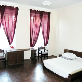 На фото: часть помещения гостиничного типа, два окна, двуспальная кровать, небольшой письменный стол, два стула, полы - паркет, стены - окрашены, на потолке - люстра