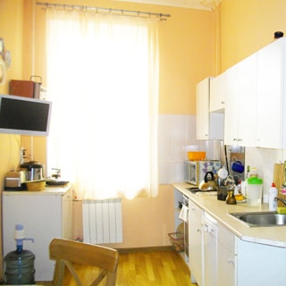 На фото: часть помещения кухни, одно окно, под окном радиатор батареи центрального отопления, слева от окна у стены - стол тумба, справа от окна вдоль стены - кухонный гарнитур с общей столешницей, столы-тумбы, кухонная плита, металлическая мойка со смесителем, навесные кухонные шкафы, фартук облицован керамической плиткой