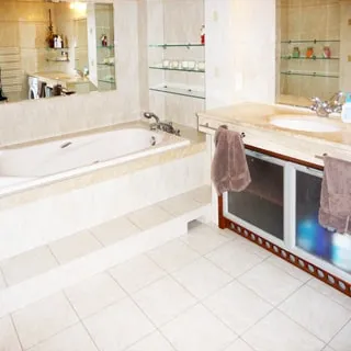 На фото: часть помещения ванной комнаты, прямо у стены - ванная, вмонтированная в основание, смеситель, зеркало, полки, справа у стены - раковина на широкой тумбе со столешницей, стены и полы облицованы плиткой