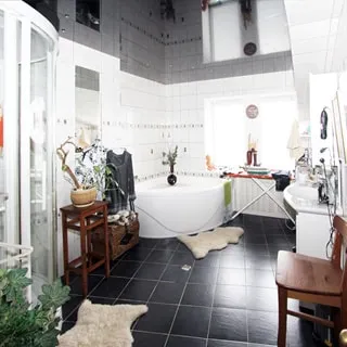 На фото: часть помещения ванной комнаты, одно окно, слева от окна угловая ванная, справа от окна у стены - керамическая раковина на тумбе, полы и стены облицованы плиткой, полы - темной, стены - белой
