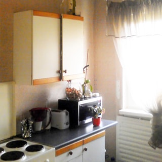 На фото: часть помещения кухни, одно окно, слева от окна вдоль стены - кухонный стол-тумба, на столе - микроволновая печь, тостер, над столом - навесной шкаф, слева от стола - четырехкомфорочная электроплита