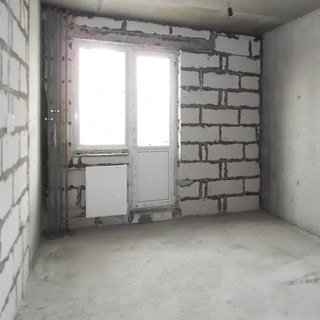 На фото: часть помещения комнаты без отделки, одно окно, установлен стеклопакет, полы - цементно-песчаная стяжка, электророзетки установлены, радиатор батареи центрального отопления отсутствует
