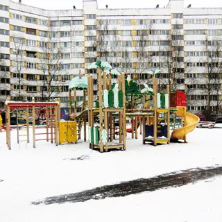 На зимнем фото: дворовая территория с оборудованной детской площадкой, на заднем плане - деревья, припаркованные у дома автомобили