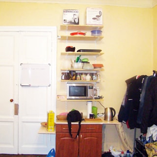 На фото: часть помещения жилой комнаты, двустворчатая входная дверь, справа от двери - кухонный стол-тумба, над ним - навесные полки, на нижней полке - микроволновая печь, справа в углу - вешалка для одежды, полы - паркет