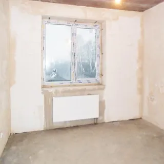На фото: помещение жилой комнаты под чистовую отделку, одно двустворчатое окно, установлен стеклопакет, под окном - батарея центрального отопления, стены оштукатурены, полы - цементно-песчаная стяжка