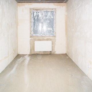 На фото: помещение жилой комнаты под чистовую отделку, одно двустворчатое окно, установлен стеклопакет, под окном - батарея центрального отопления, стены оштукатурены, полы - цементно-песчаная стяжка