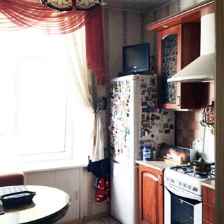 На фото: часть помещения кухни, окно, слева от окна - обеденный стол и кухонный мягкий уголок, справа от окна вдоль стены - двухкамерный холодильник, на нем - небольшой телевизор, кухонная тумба-стол, над ней - навесной кухонный шкаф, правее - четырехкомфорочная газовая плита с духовым шкафом, над ней - вытяжка, правее - еще одна кухонная тумба-стол