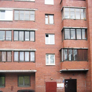На фото: часть фасада кирпичного многоэтажного многоквартирного жилого дома, балконы застеклены, на входе в парадную - металлические двери, домофон, на окнах первого этажа - защитные решетки