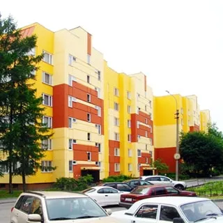 На фото: часть фасада панельного шестиэтажного многоквартирного жилого дома, фасад раскрашен в три цвета (оранжевый, ярко-желтый и песочный), без балконов, благоустроенная придомовая территория, газоны, деревья, тротуар, проезжая часть, припаркованные автомобили