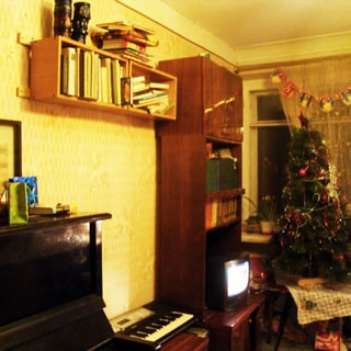 На фото: часть помещения жилой комнаты, окно, у окна столик с новогодней елкой, слева от окна вдоль стены - мебельная секция с полками для книг и тумбой под телевизор, леве на стене - книжная полка, левее у стены - пианино, стены оклеены обоями
