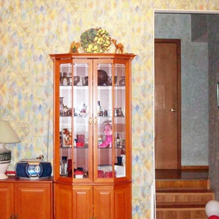 На фото: часть помещения жилой комнаты - гостиной, в центре у стены - сервант, слева от него - тумба с магнитолой и настольной лампой, справа - арка и ступеньки вверх в прихожую, стены оклеены обоями