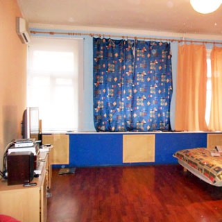 На фото: часть помещения жилой комнаты, три окна, слева у стены тумба с телевизором, над ней на стене - кондиционер, справа - часть разложенного дивана или кровати, полы - ламинат, на потолке - люстра