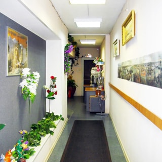 На фото: часть нежилого помещения - коридора или холла, стены окрашены, украшены эстампами и цветами, на полу - ковровая дорожка, на потолке - свтельники дневного света, на дальнем плане - входной холл и входная дверь