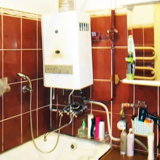 На фото: часть помещения ванной комнаты, прямо у стены в углу - ванная со смесителем, над ней - газовая колонка, правее на стене - шкафчик с зеркальными дверцами, стены облицованы керамической плиткой