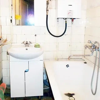 На фото: часть помещения ванной комнаты, прямо у стены в углу на тумбе белого цвета с дверцами - керамическая раковина со смесителем, над раковиной - зеркало, справа - ванная со смесителем, над ванной - газовая колонка, стены в половину высоты облицованы керамической плиткой
