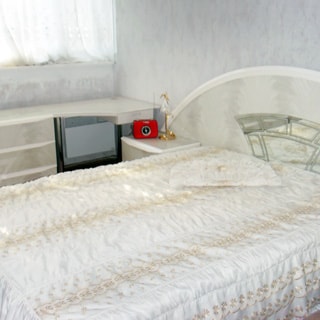 На фото: часть помещения жилой комнаты - спальни, окно, у окна - комод, туалетный столик, у стены - двуспальная кровать, слева от кровати - прикроватная тумбочка с ночником и будильником