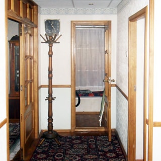 На фото: часть помещения прихожей, в центре, слева и справа - двери в соседние помещения, слева в углу вешалка - стойка для верхней одежды, над ней на стене - часы, стены оклеены обоями, на полу - ковер