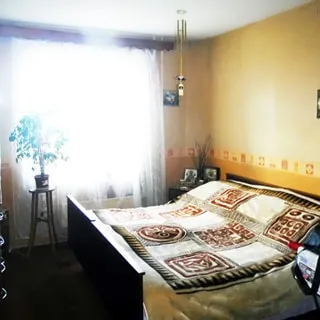 На фото: часть помещения жилой комнаты - спальни, двустворчатое окно, у стены - двуспальная кровать, слева от кровати - прикроватная тумбочка, у окна - высокая цветочная стойка, стены оклеены обоями