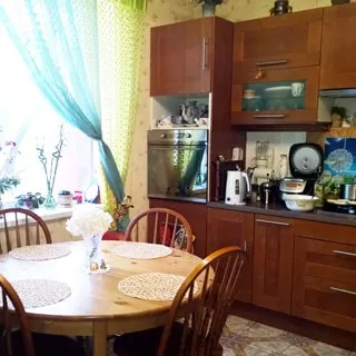 На фото: часть помещения кухни-столовой, окно, справа от окна вдоль стены - кухонный гарнитур со встроенной кухонной техникой, кухонная посуда, слева у окна - круглый обеденный стол со стульями, полы - плитка