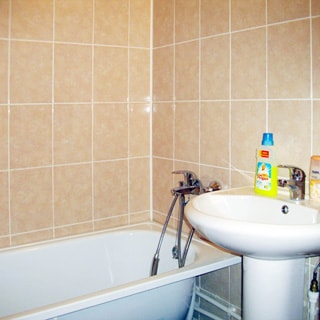 На фото: часть помещения ванной комнаты, керамическая раковина на стойке, со смесителем, слева от раковины - ванная со смесителем, стены облицованы керамической плиткой бежевого оттенка