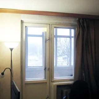 На фото: часть помещения жилой комнаты, одно с балконной дверью, за окном - застекленная лоджия, под окном - радиатор центрального отопления, слева от окна - торшер