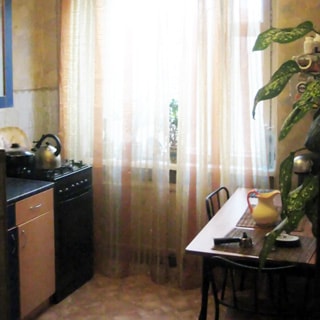 На фото: часть помещения кухни, большое окно, слева от окна - 4-комфорочная газовая плита с духовкой, кухонная тумба-стол, под окном - радиатор центрального отопления, справа от окна - обеденный стол, два стула у стола, полы - линолеум