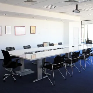 На фото: часть офисного помещения для проведения совещаний, большой прямоугольный стол, офисные стулья и кресло руководителя, потолки - навесные с встроенными светильниками, на потолке установлен проектор, полы - ковролин синего цвета