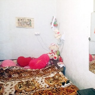 На фото: часть помещения жилой комнаты, прямо у стены диван-кровать в разложенном состоянии застелен пледом, справа на стене - лампа для чтения, зеркала, стены оклеены светлыми обоями