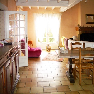 На фото: часть помещения гостиной с выходом в кухню и на террасу, слева - комод, у выхода на террасу - мягкое кресло, справа - стол со стульями, на дальнем плане - камин, мягкие кресла, полы - плитка