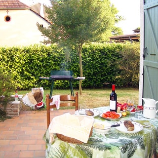 На фото: мощеная плиткой терраса с накрытым обеденным столом, на заднем плане - дымящийся мангал, газон, живая изгородь из кустов