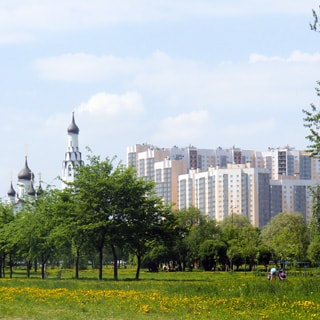 На фото: территория парка или сквера, газоны, деревья, на дальнем плане - православный храм, еще дальше - многоэтажные жилые дома